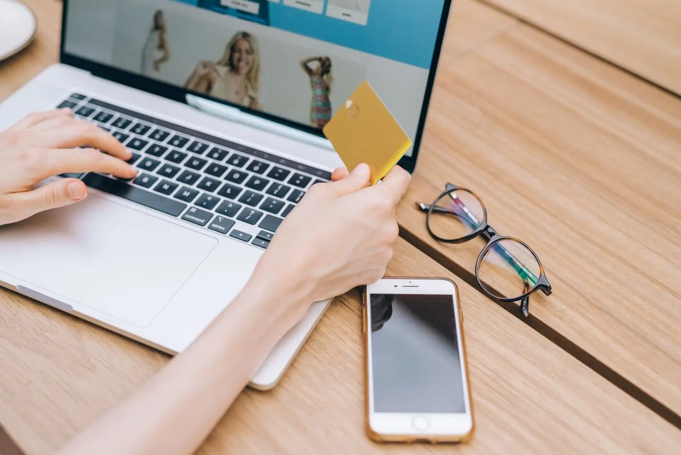 Person zahlt online mit Kreditkarte am Laptop, Smartphone und Brille liegen daneben